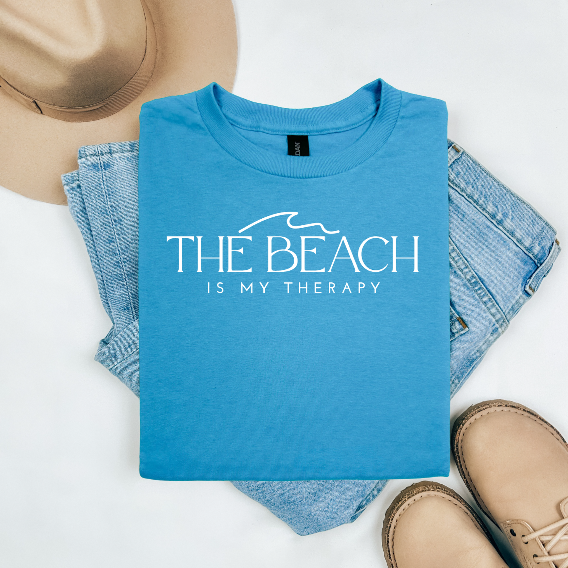 Gildan 5000 "The Beach Is My Therapy" t shirt with a simple wave graphic in one color. Picture on a flay lay mockup with pair of blue jeans, brown shoes and a hat accessory.
