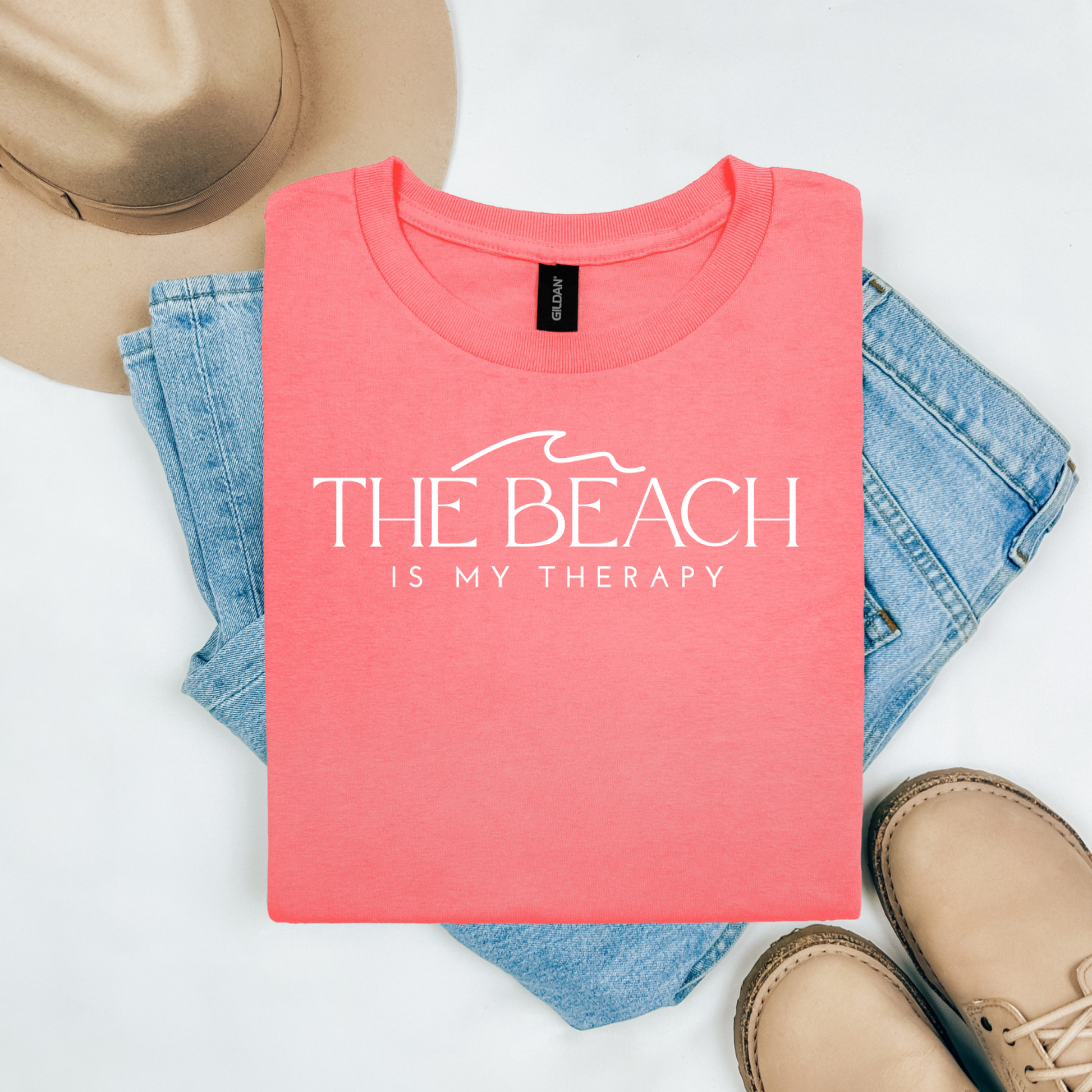 Gildan 5000 "The Beach Is My Therapy" t shirt with a simple wave graphic in one color. Picture on a flay lay mockup with pair of blue jeans, brown shoes and a hat accessory.