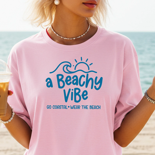 Person wearing a pink t-shirt with 'a Beachy Vibe' text on a beach.