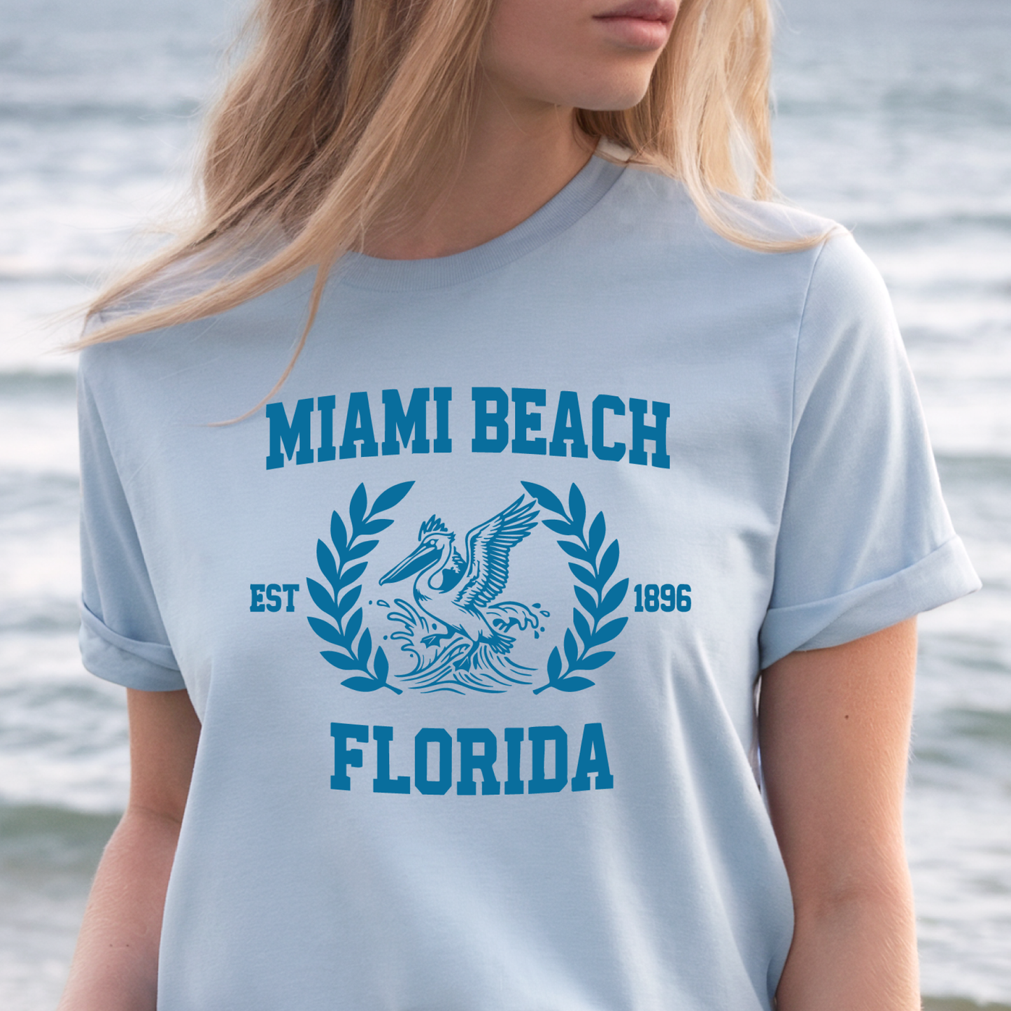 Person wearing a light blue t-shirt with 'Miami Beach Florida' design by the water.