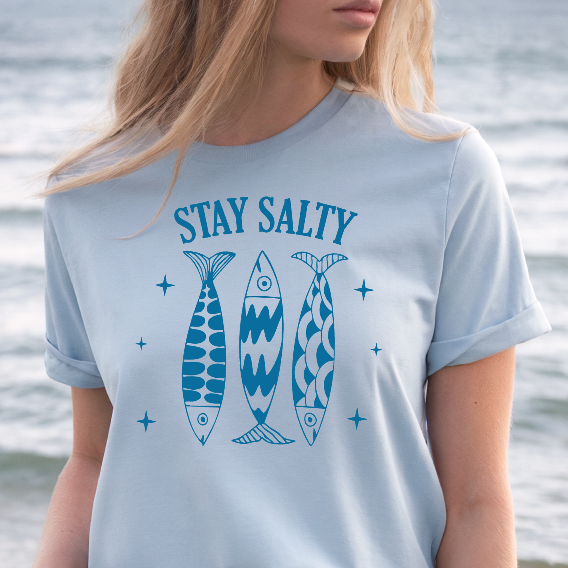 Person wearing a light blue t-shirt with 'Stay Salty' text and fish graphics, standing by water.
