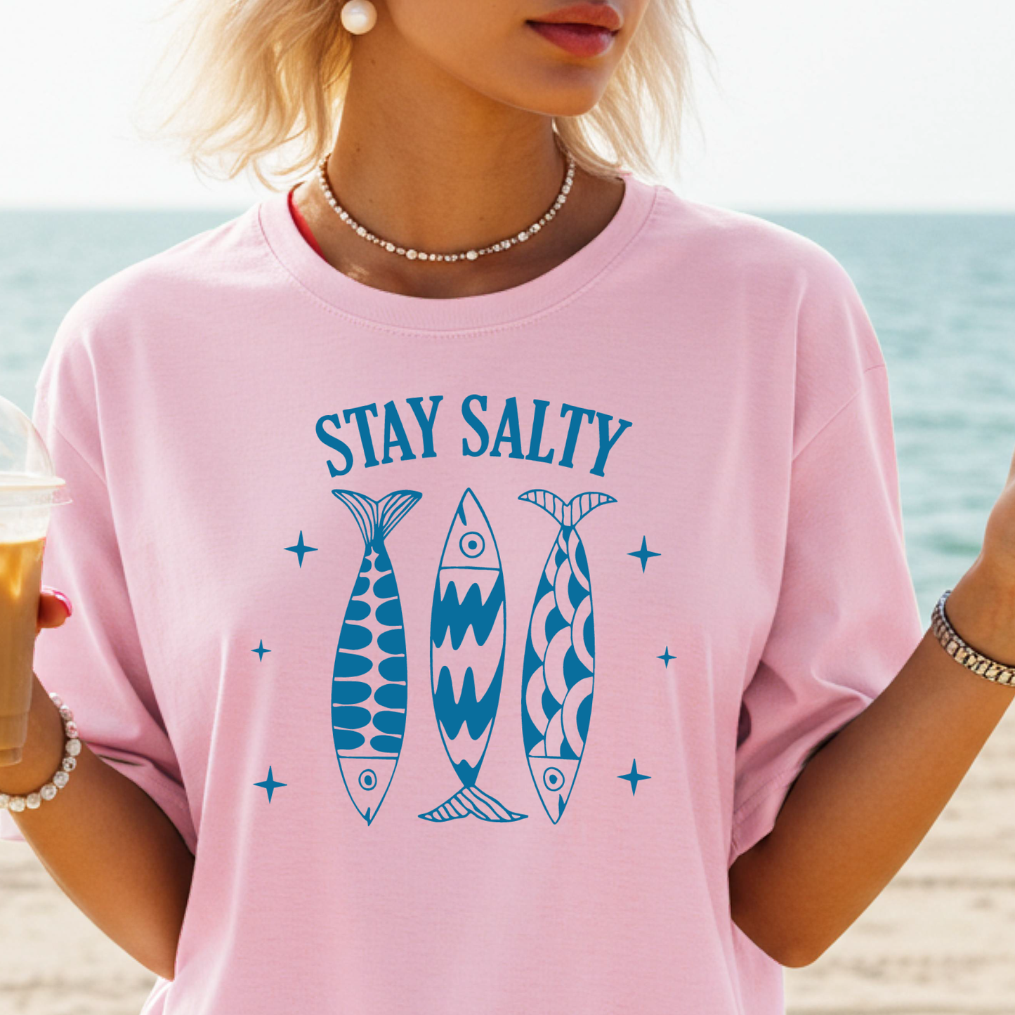 Person wearing a pink t-shirt with 'Stay Salty' and fish graphics on a beach.
