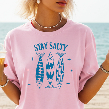 Person wearing a pink t-shirt with 'Stay Salty' and fish graphics on a beach.