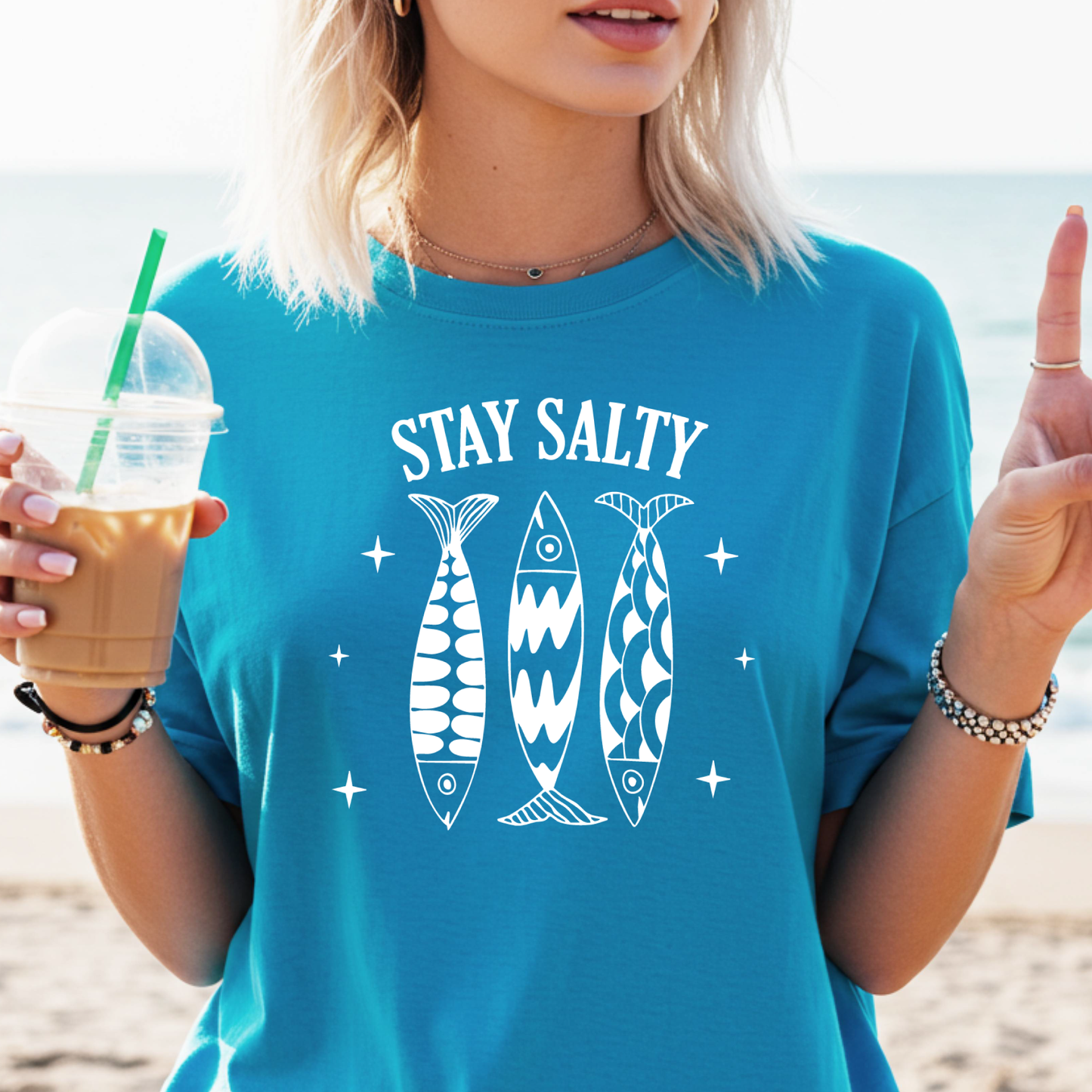 Person wearing a sapphire blue t-shirt with 'Stay Salty' and fish graphics, holding a drink on a beach.