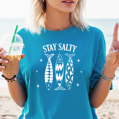 Person wearing a sapphire blue t-shirt with 'Stay Salty' and fish graphics, holding a drink on a beach.
