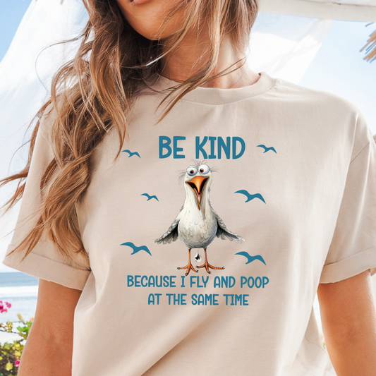 Person wearing a natural t-shirt with a cartoon seagull and text 'Be kind because I fly and poop at the same time'.