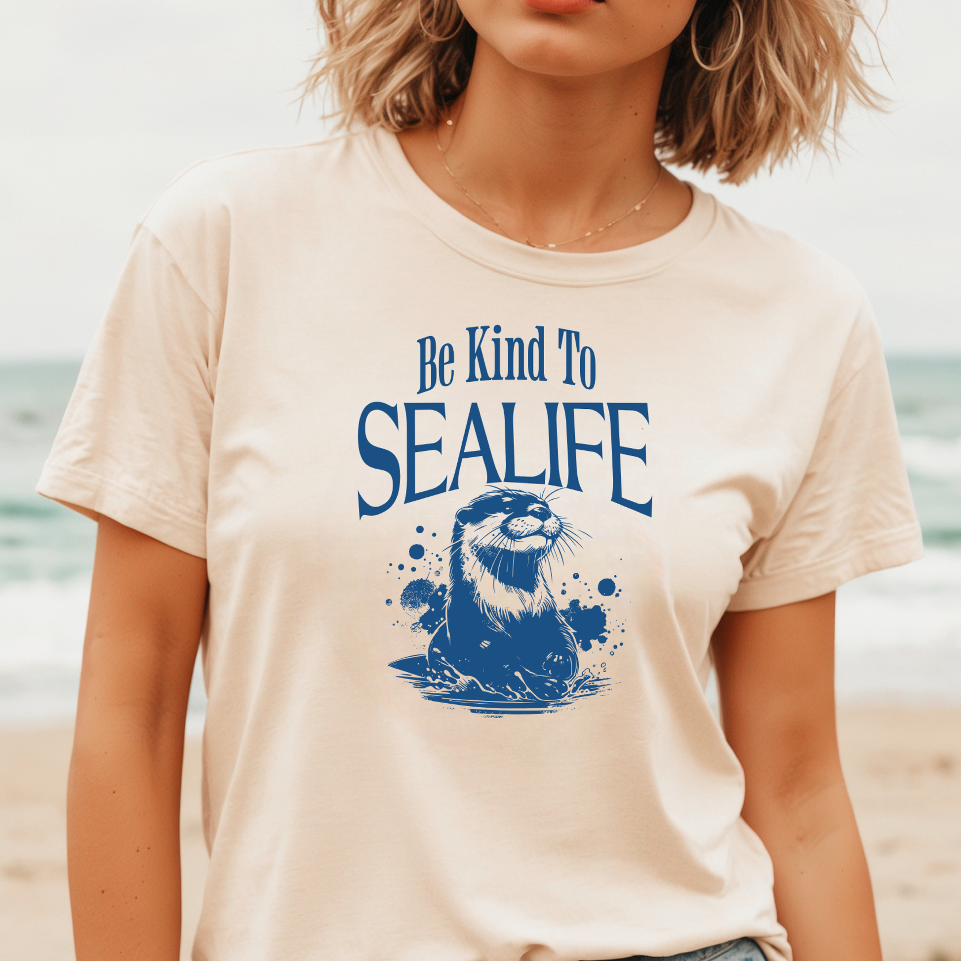 Gildan 5000 "Be Kind To Sealife" t shirt with a otter splashing in the water in a one color graphic. Picture of a girl standing on the beach in a t shirt.