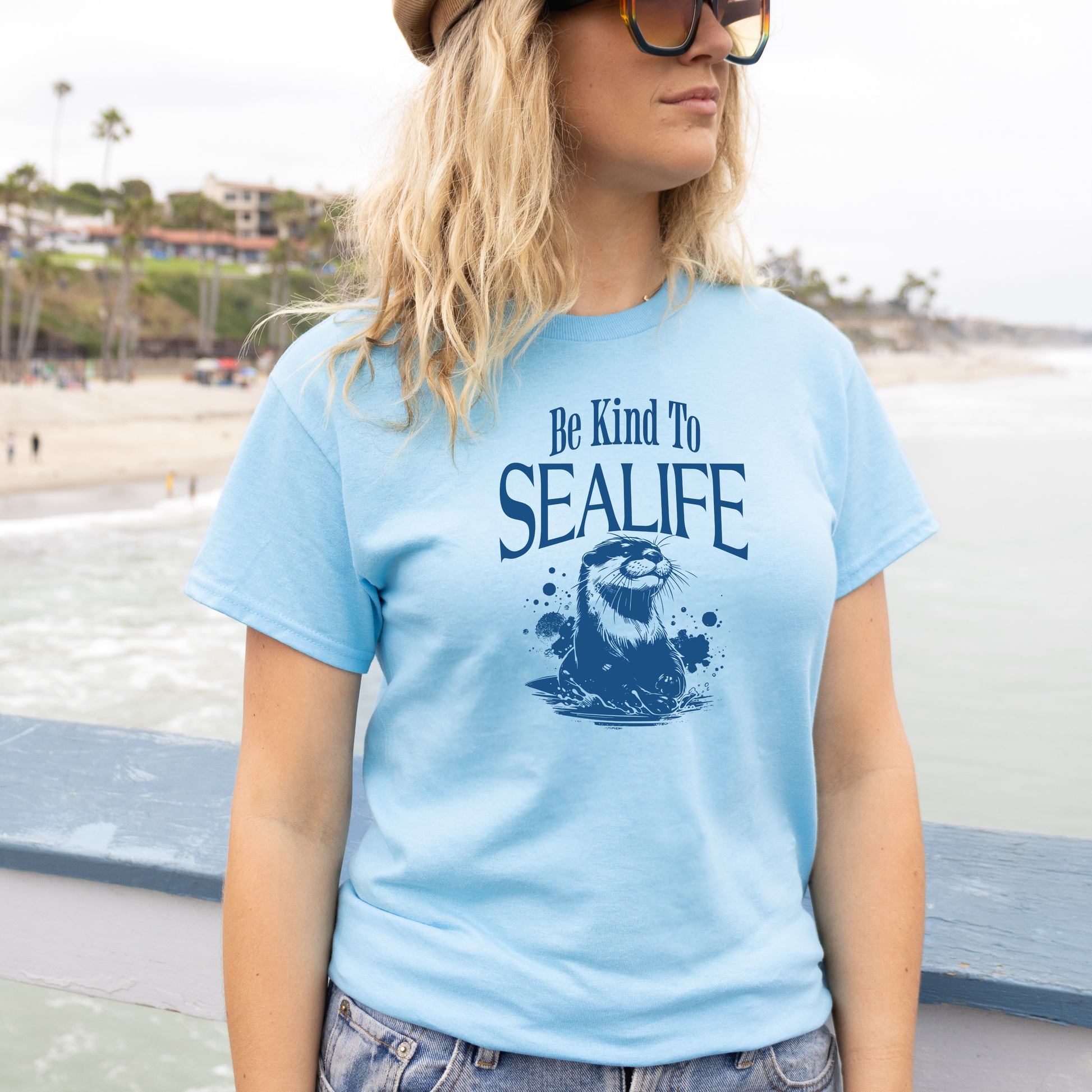 Gildan 5000 "Be Kind To Sealife" t shirt with a otter splashing in the water in a one color graphic. Picture of a girl standing on the beach in a t shirt.