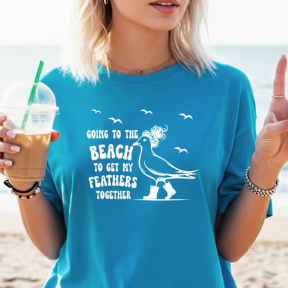 Person wearing a sapphire blue t-shirt with a beach-themed graphic and text, holding a drink on a beach.