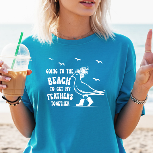 Person wearing a sapphire blue t-shirt with a beach-themed graphic and text, holding a drink on a beach.