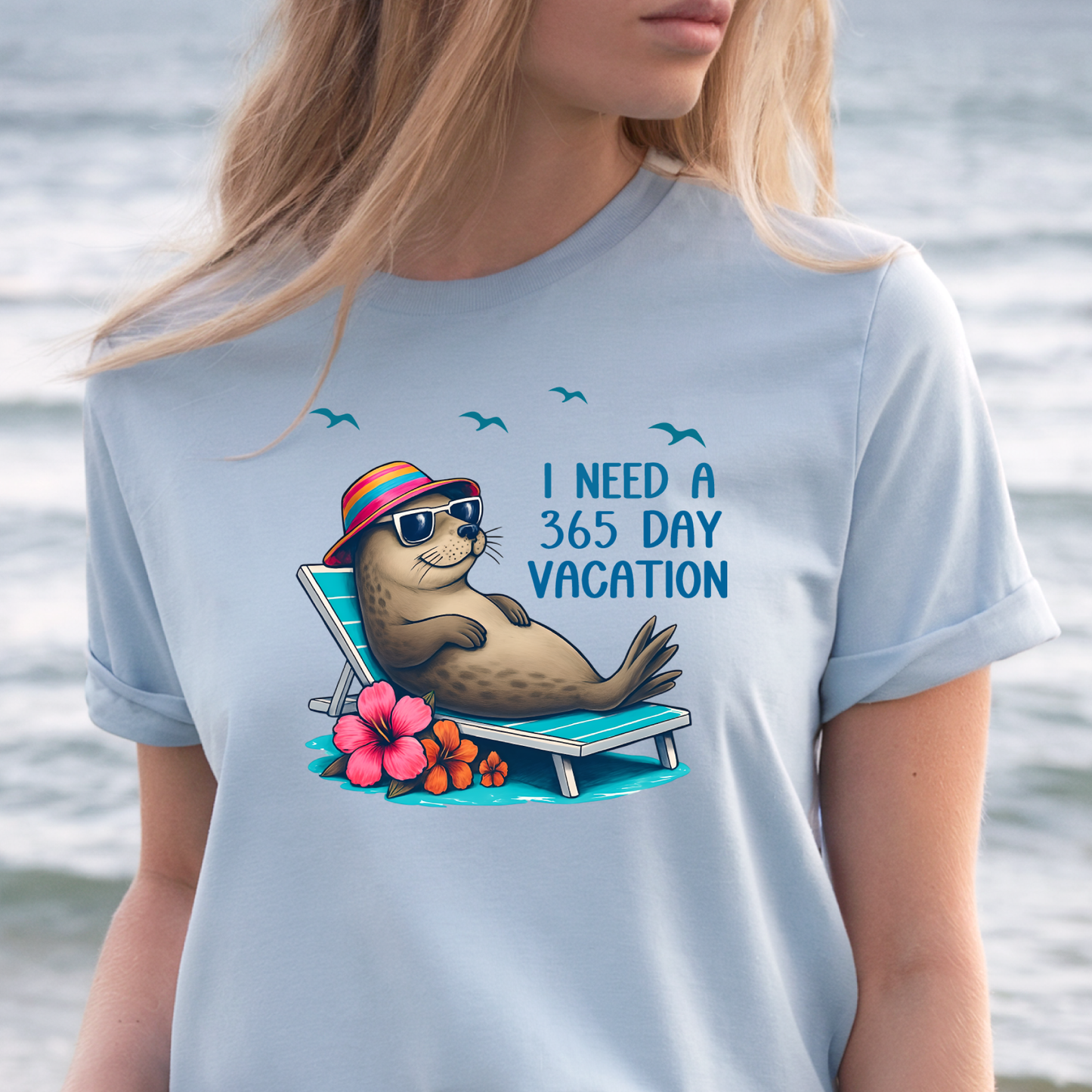 Person wearing a light blue t-shirt with a seal graphic and text by a body of water.