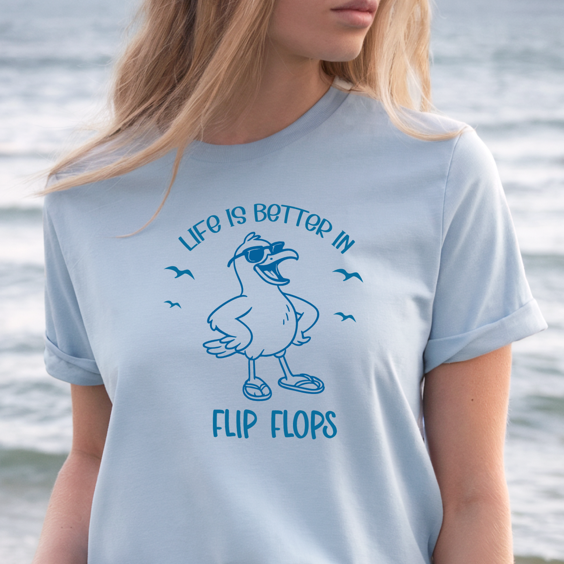 Person wearing a light blue t-shirt with a graphic and text 'Life is better in flip flops' against a water background.