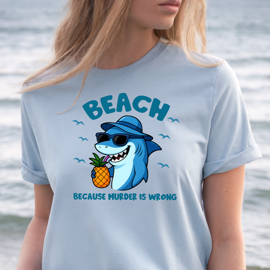 Person wearing a light blue t-shirt with a shark graphic and text by the water.