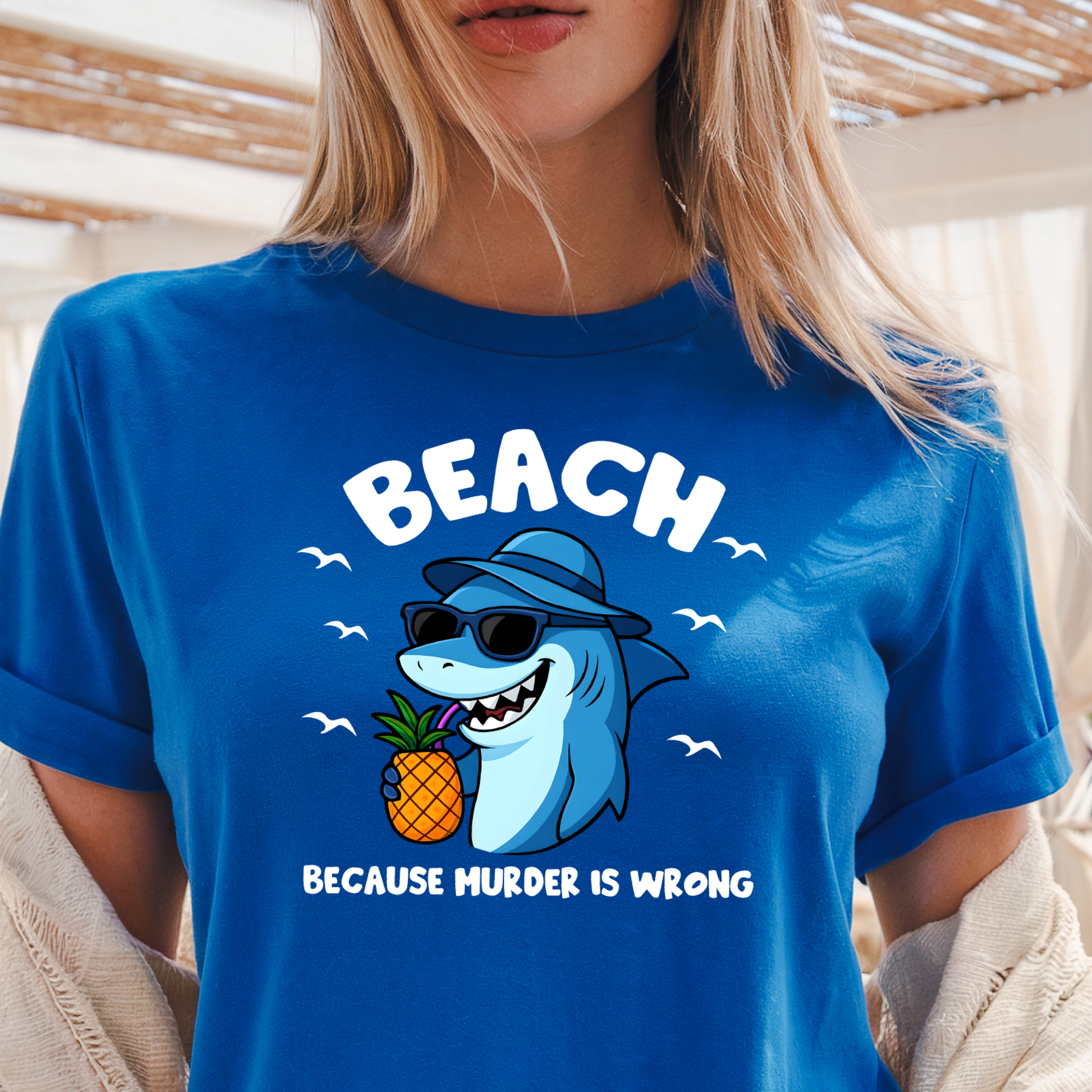 Blue t-shirt with a shark holding a pineapple and text 'BEACH BECAUSE MURDER IS WRONG' worn by a person.