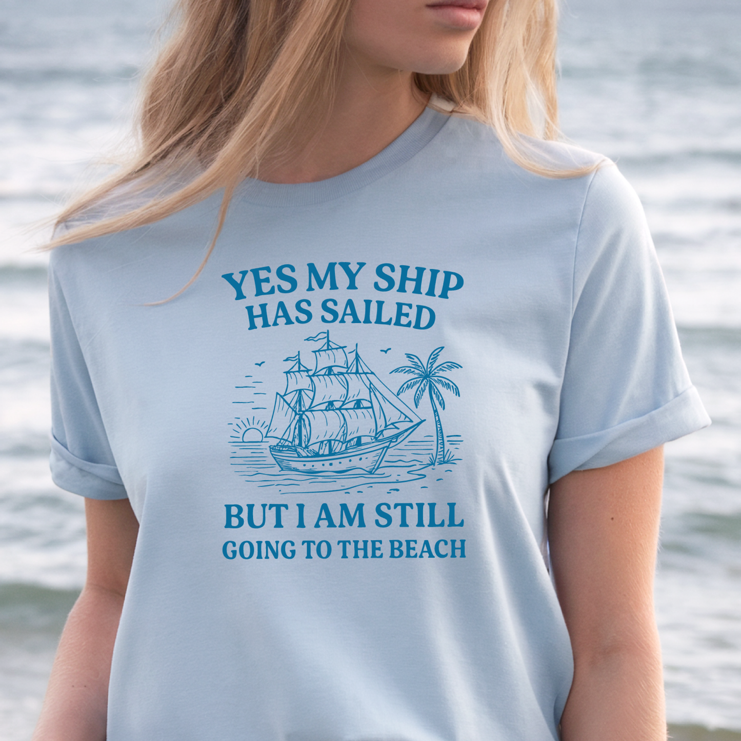 Person wearing a light blue t-shirt with a ship and beach graphic and text by the water.