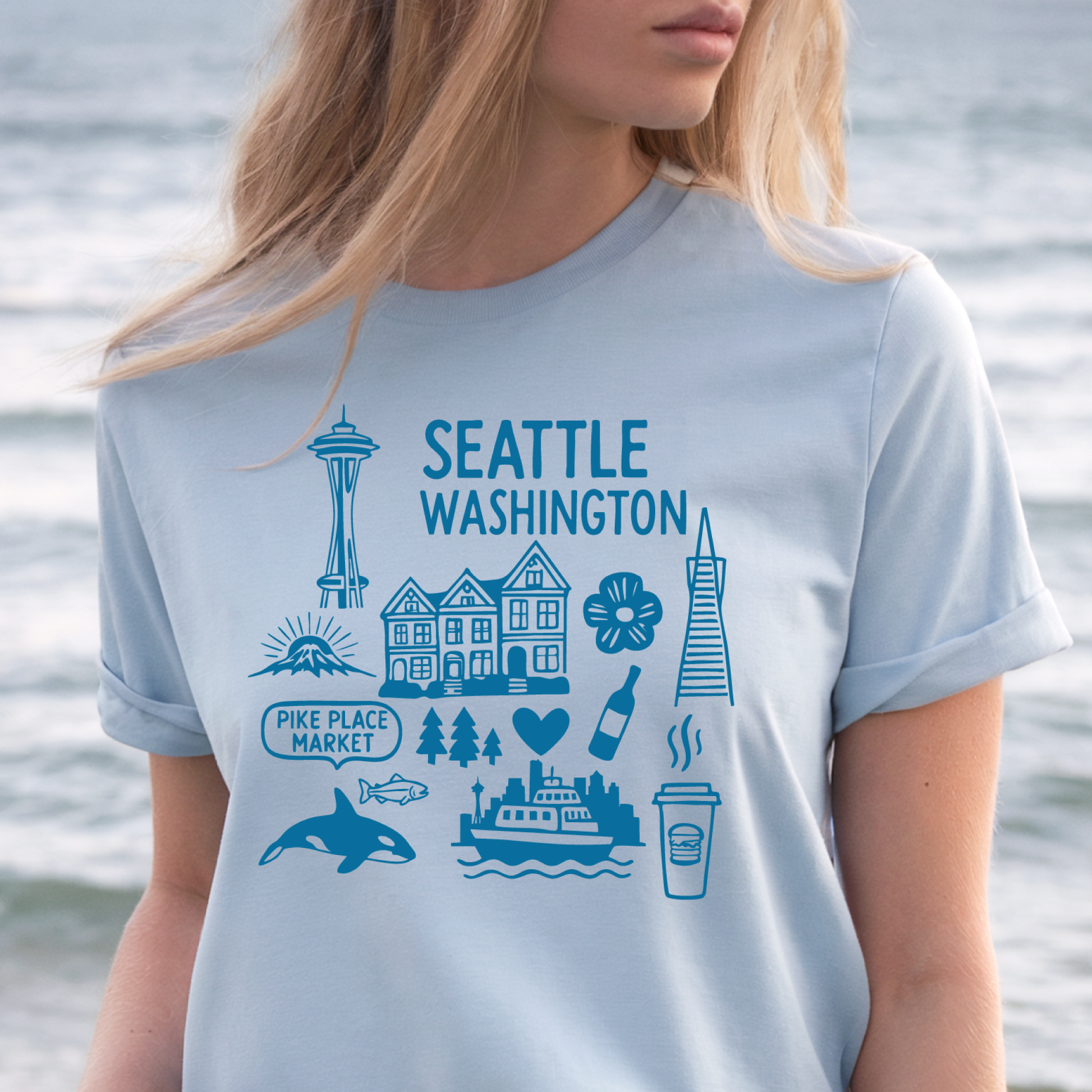 Person wearing a light blue t-shirt with Seattle-themed graphics against a water background