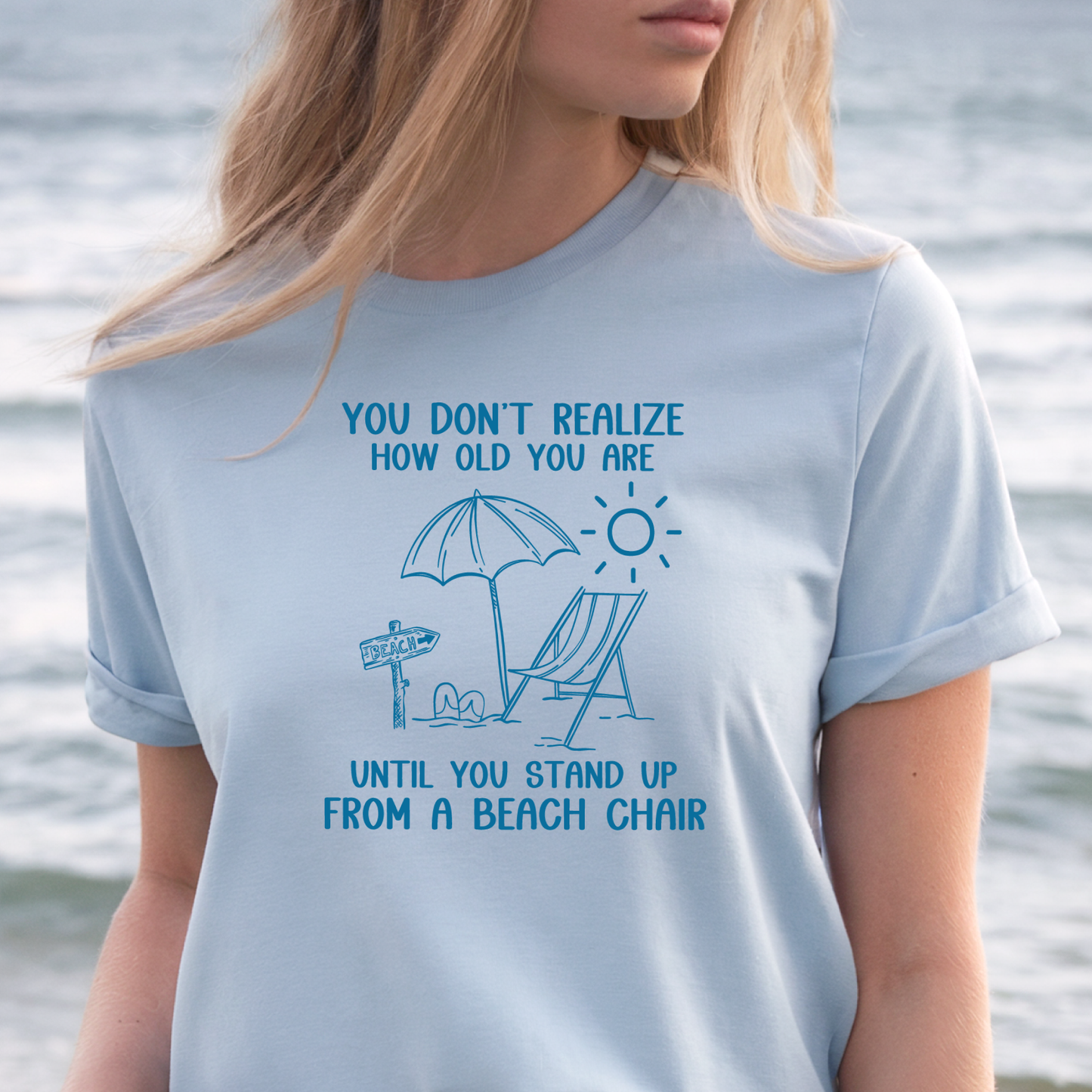 Person wearing a light blue t-shirt with a beach-themed design and text, standing by water.
