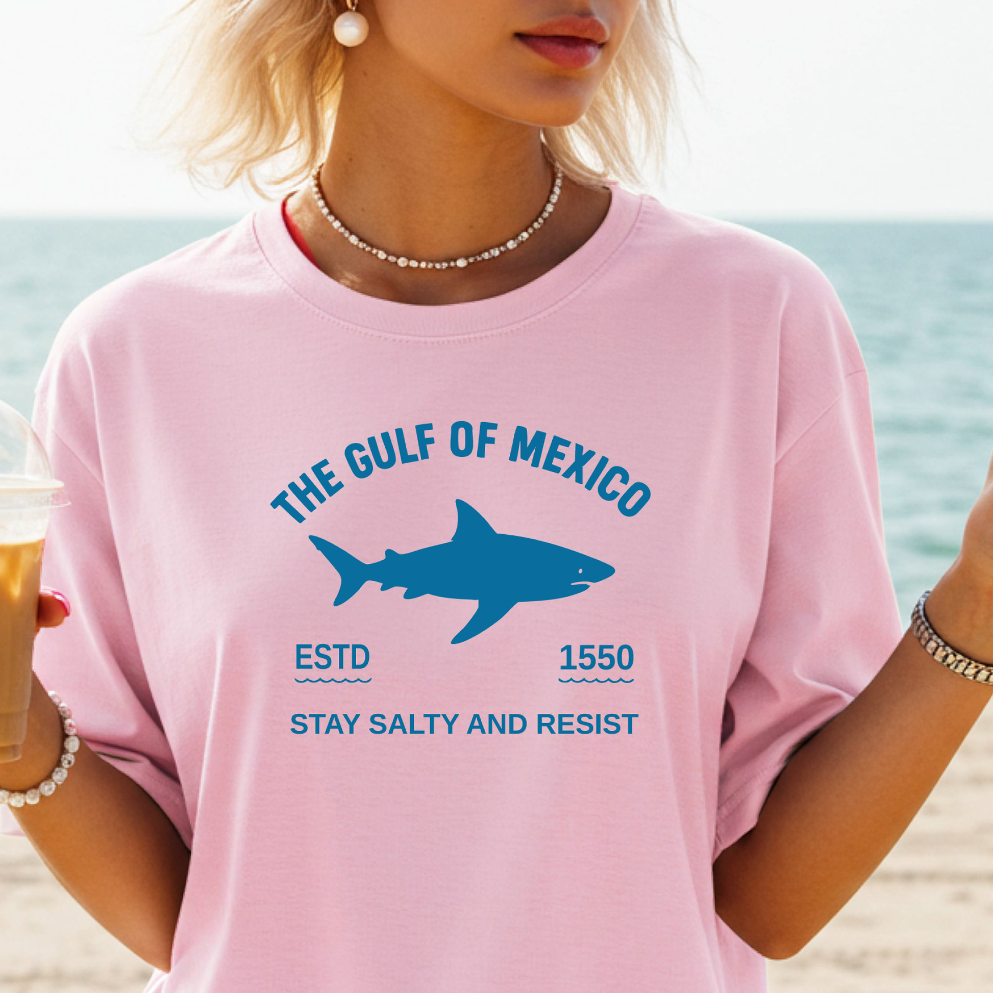 Person wearing a pink t-shirt with a shark graphic and text on a beach.