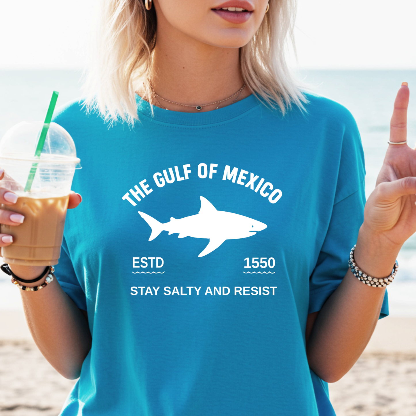 Person wearing a sapphire blue t-shirt with a shark graphic and text on a beach.