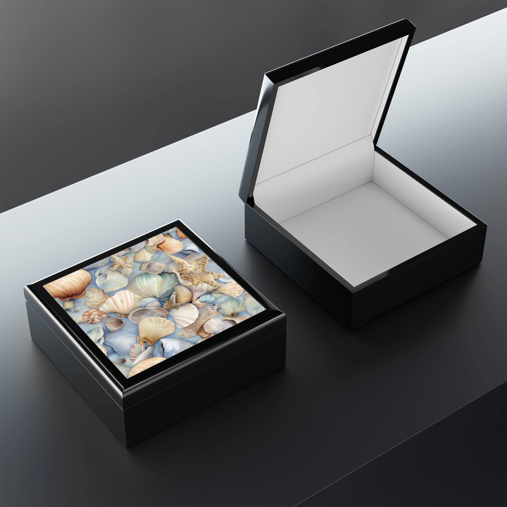 Two black square boxes serve as perfect nautical-themed decor. One boxs lid features a seashell pattern, while the other reveals a plain white interior. Ideal as elegant bridal gifts, these Coastal Shells Jewelry Boxes rest on a reflective gray surface.