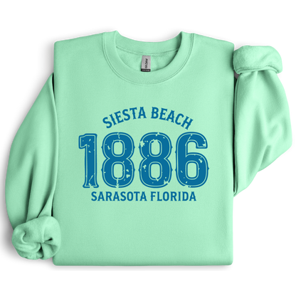 Light green sweatshirt with blue text 'Siesta Beach 1886 Sarasota Florida' on a white background