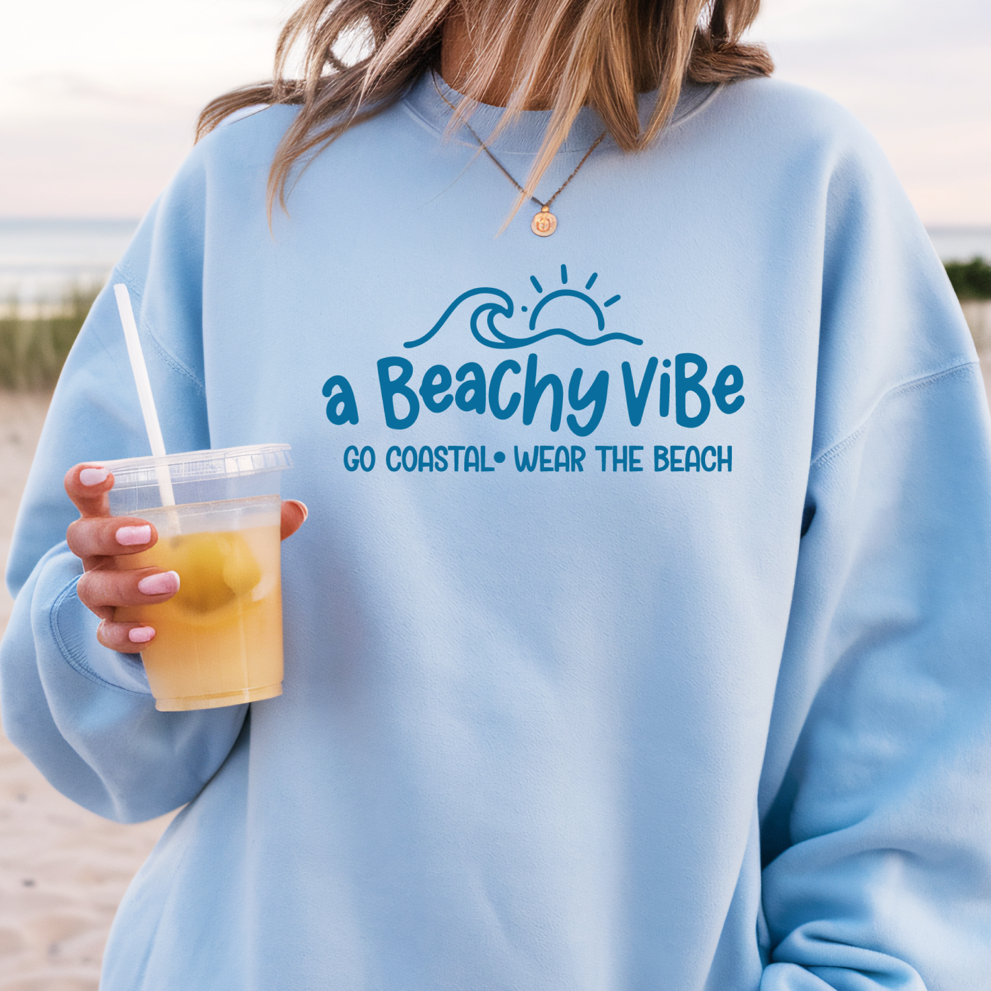 Person wearing a light blue sweatshirt with 'a Beachy Vibe' text and graphics, holding a drink on a beach.