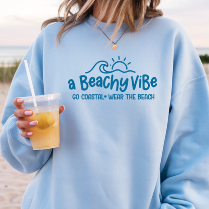 Person wearing a light blue sweatshirt with 'a Beachy Vibe' text and graphics, holding a drink on a beach.