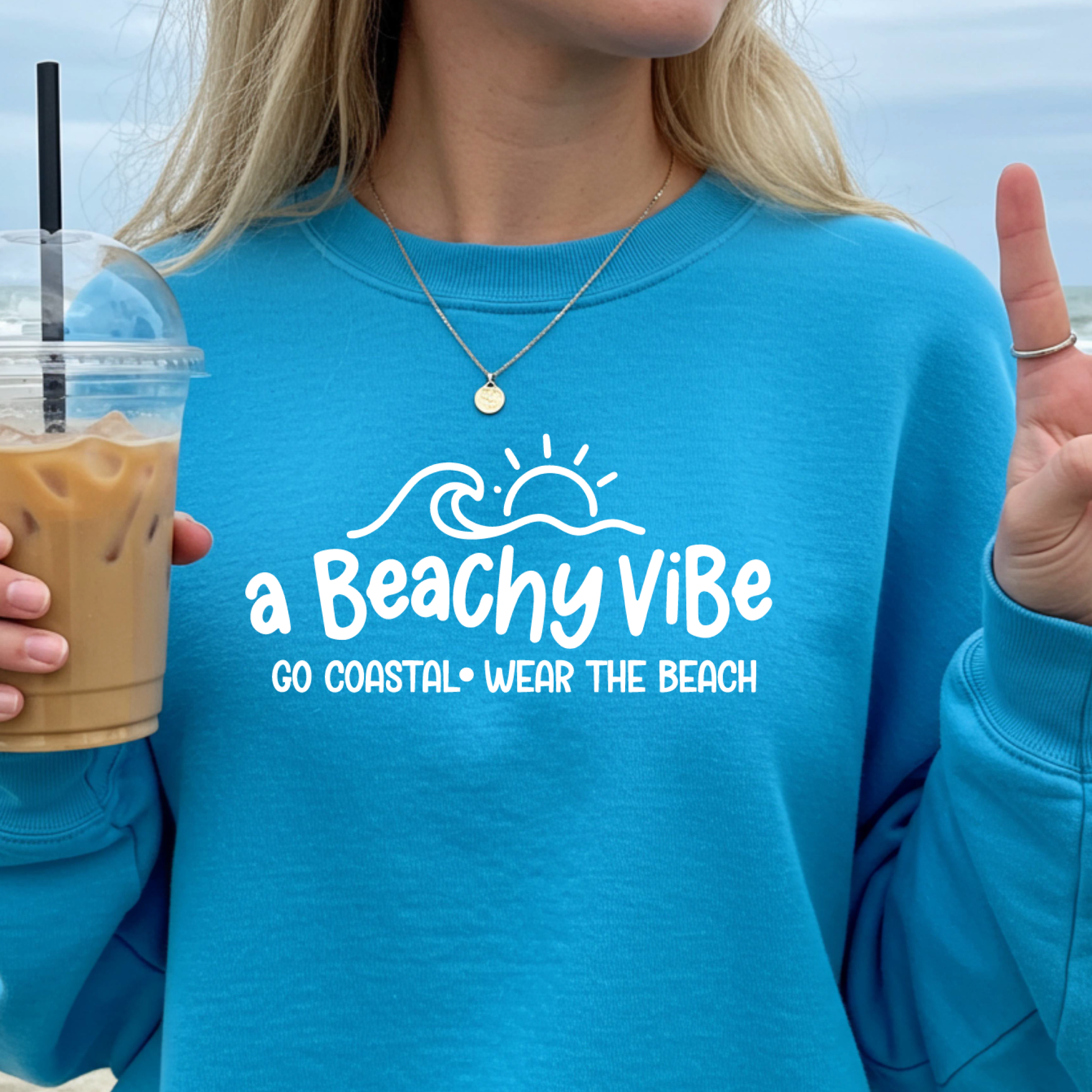 Person wearing a sapphire blue sweatshirt with 'a Beachy Vibe' text, holding a drink.