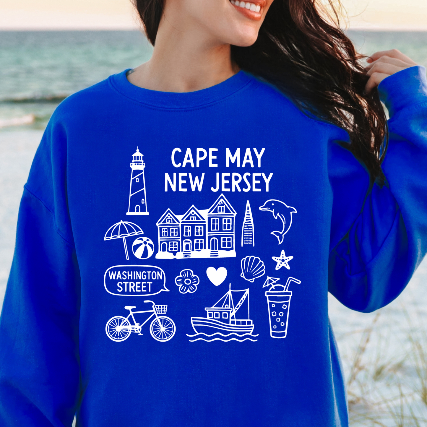 Gildan sweatshirt with blue Cape May New Jersey design featuring beach icons like a lighthouse, dolphin, beach ball, shells, and Washington Street sign. Coastal-inspired pullover made from soft cotton-poly blend fabric, ideal for beach lovers and Cape May fans. Perfect for seaside getaways or relaxing at home, this cozy crewneck celebrates the charm of New Jersey’s iconic coastal town.