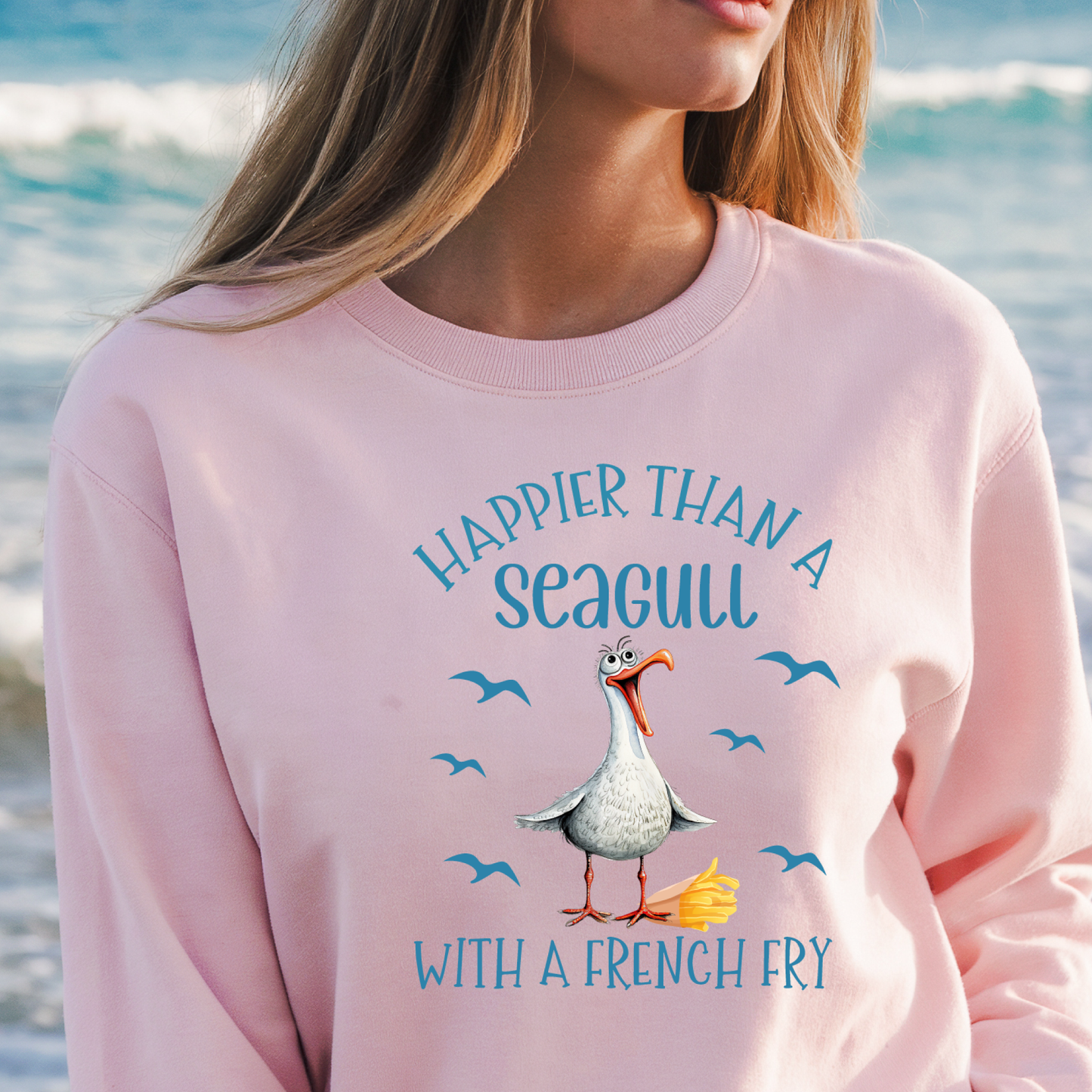 Person wearing a pink sweatshirt with a seagull graphic and text by the ocean.