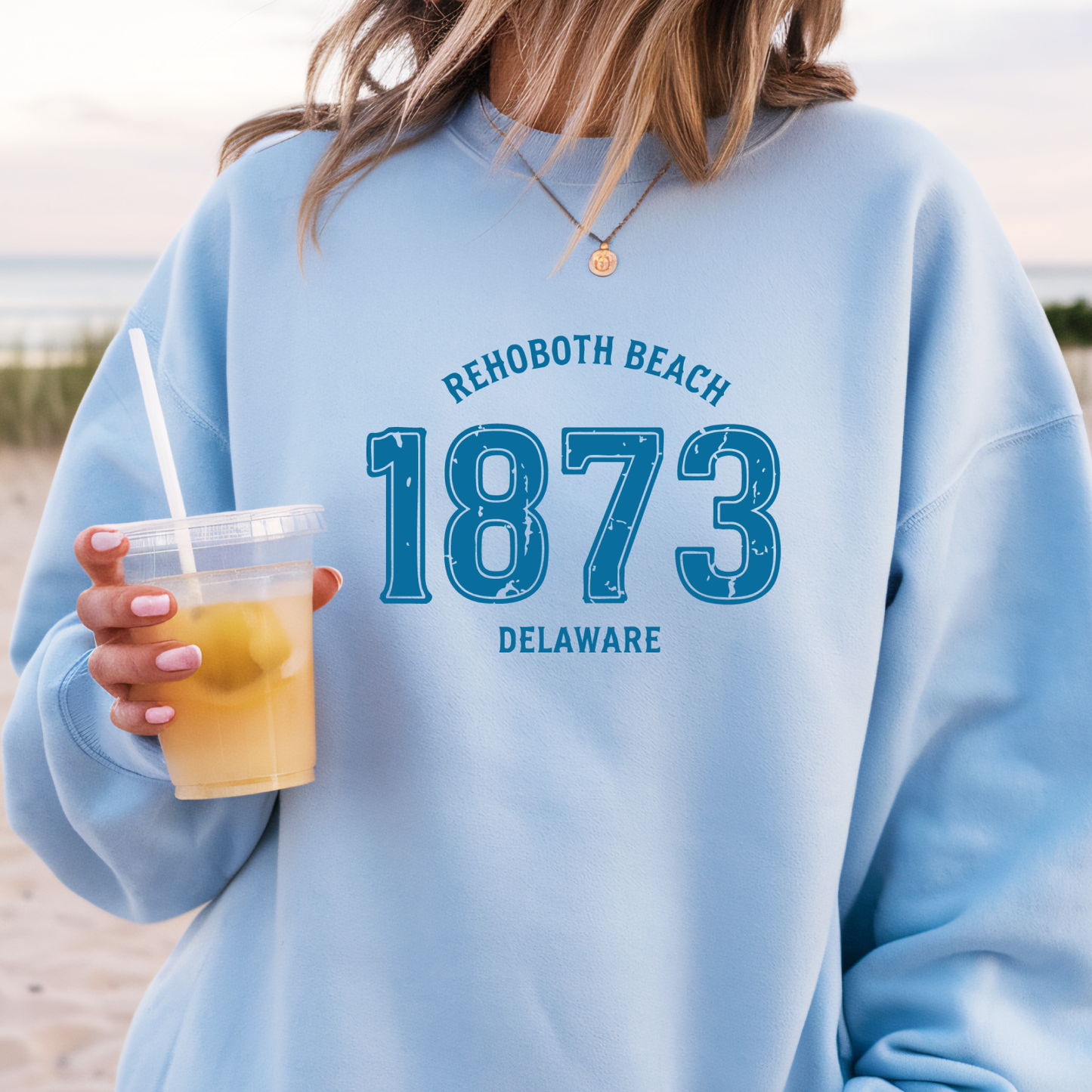 Person wearing a light blue sweatshirt with 'Rehoboth Beach 1873 Delaware' text, holding a drink on a beach.