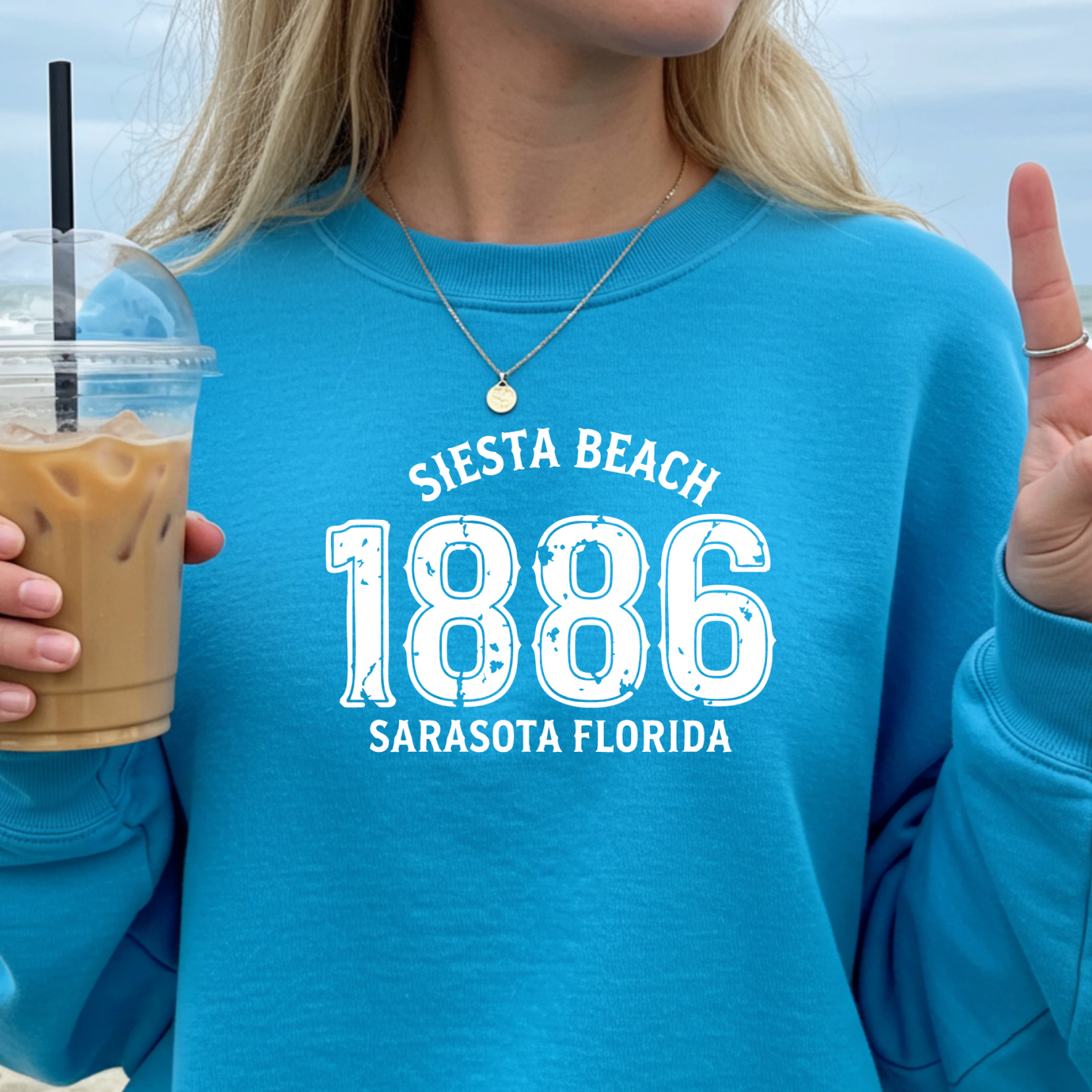 Person wearing a sapphire blue sweatshirt with 'Siesta Beach 1886 Sarasota Florida' text, holding a drink.