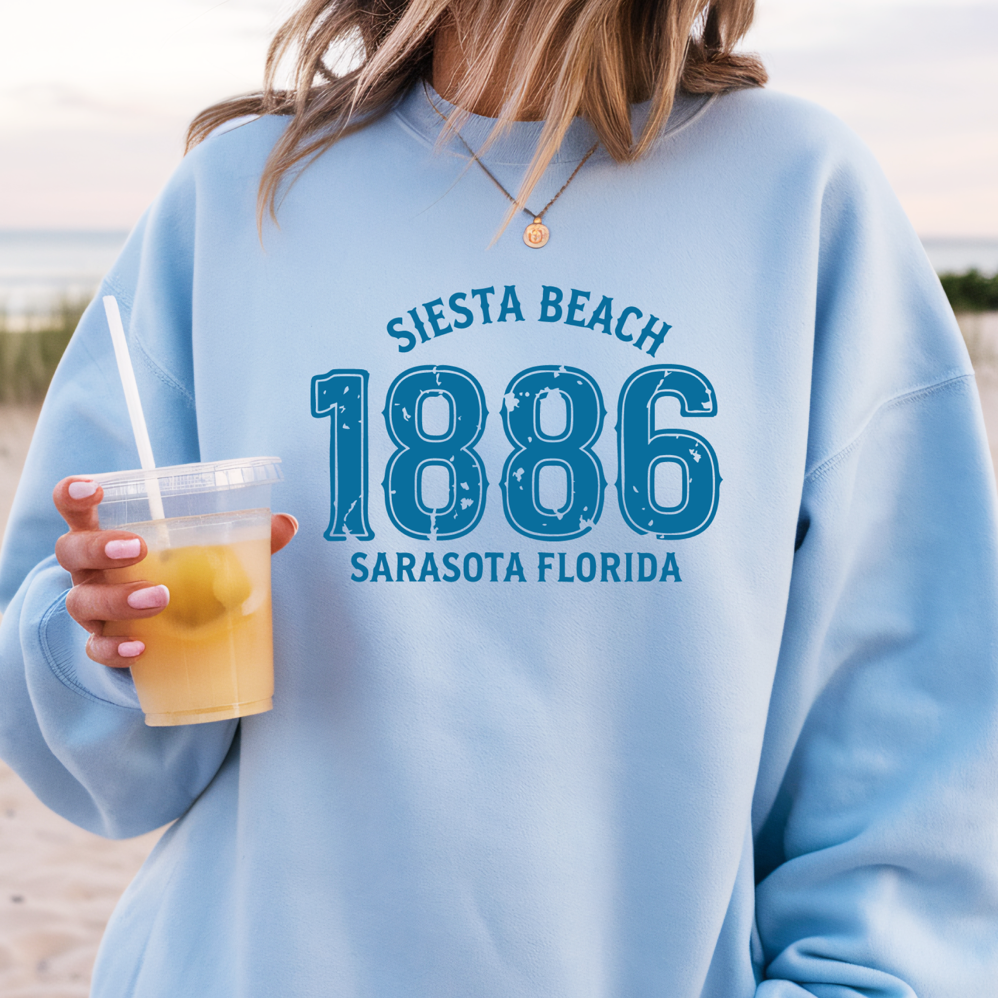 Person wearing a light blue sweatshirt with 'Siesta Beach 1886 Sarasota Florida' text, holding a drink on a beach.
